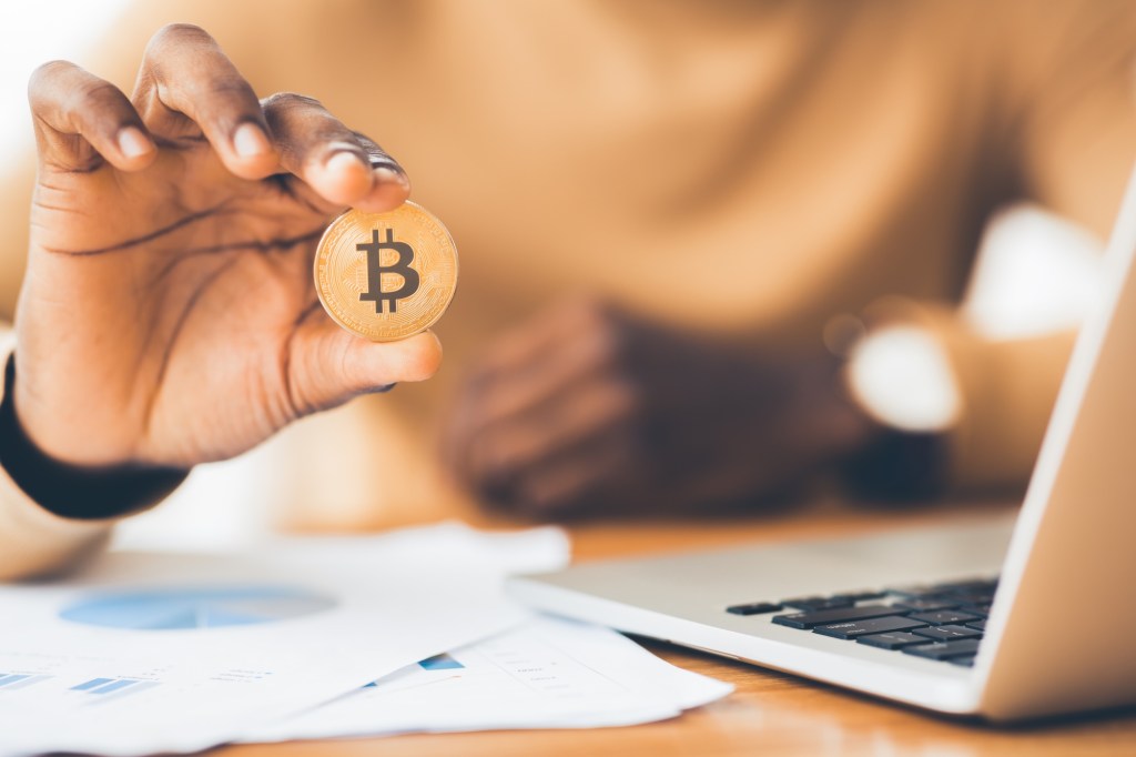 Closeup on hand holding Bitcoin coin near laptop screen on the table.
