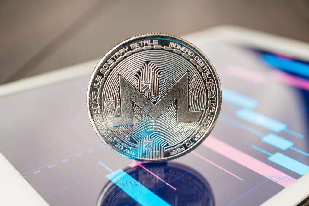 A shiny, silver Monero coin sits centered on a digital tablet displaying colorful financial data.