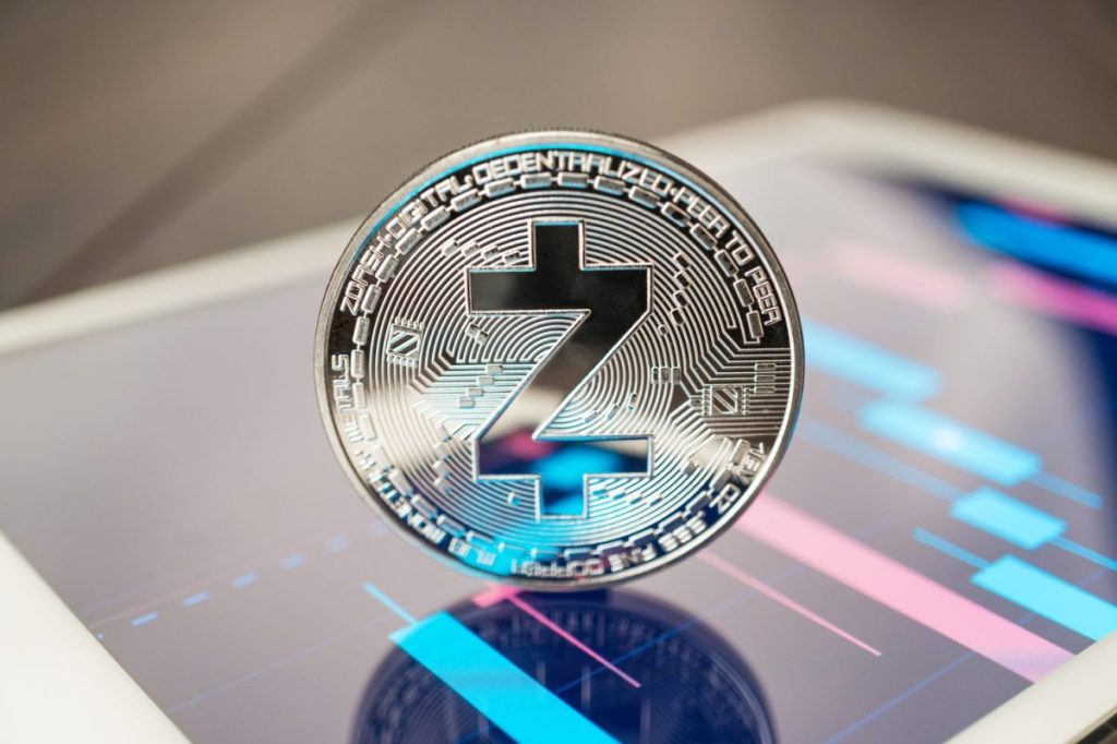 Close-up captures a shiny cryptocurrency Zcash coin. The metallic coin rests on a digital tablet. The tablet displays digital graph lines in shades of blue and pink. The coin reflects these colors.