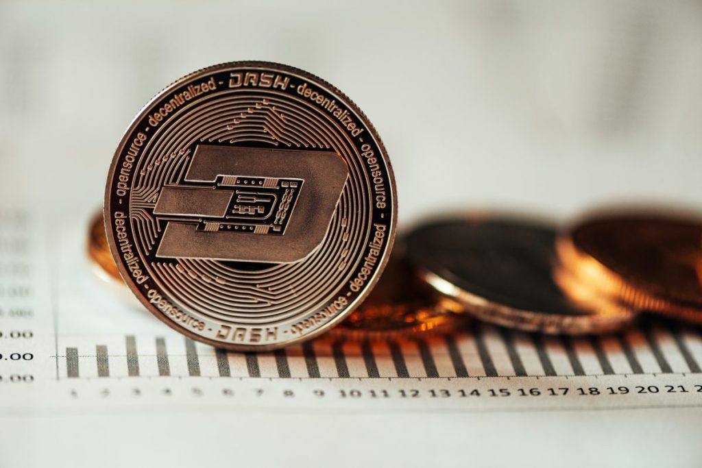 A single dash cryptocurrency coin sits atop a financial chart in this close-up shot. The coin is copper-colored with a black inlay, and the words "dash - decentralized - opensource" are inscribed around the edge. The chart beneath it has a series of bars and numbers.